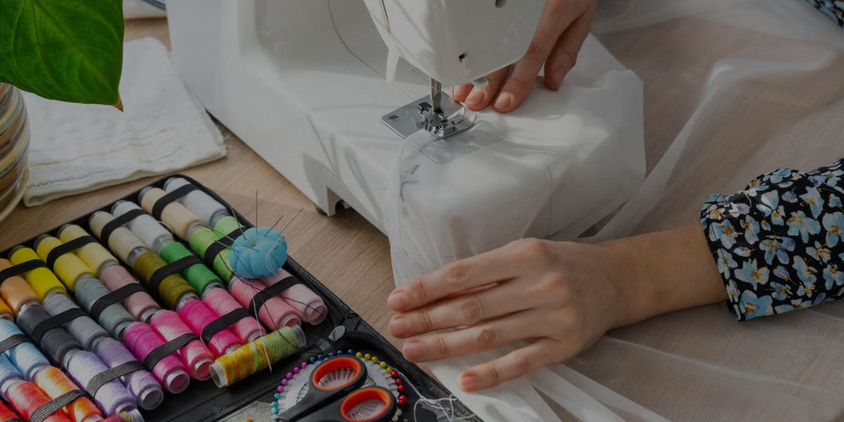 A close-up of a sewing machine in use with white fabric, showing a person's hands guiding the material. A tray of colorful sewing thread spools and scissors sits on the table in the foreground.