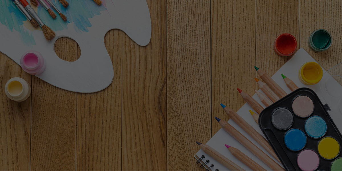 A wooden tabletop styled with art supplies, including a white paint palette with paintbrushes, small paint pots, colored pencils, and a watercolor paint palette on a notebook.