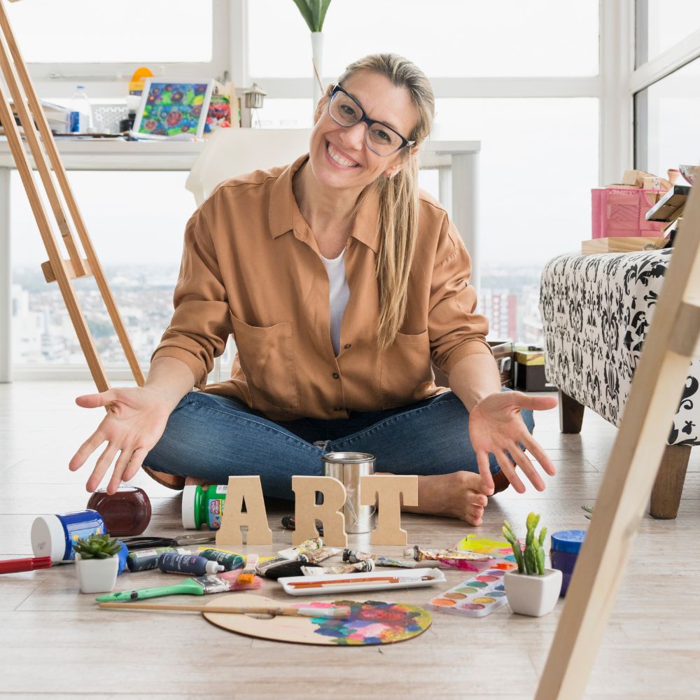 A smiling woman with glasses sits cross-legged on a wooden floor in a bright room, surrounded by art supplies and wooden letters spelling ART. She wears a brown button-up shirt and blue jeans, with easels and a patterned sofa visible in the background.