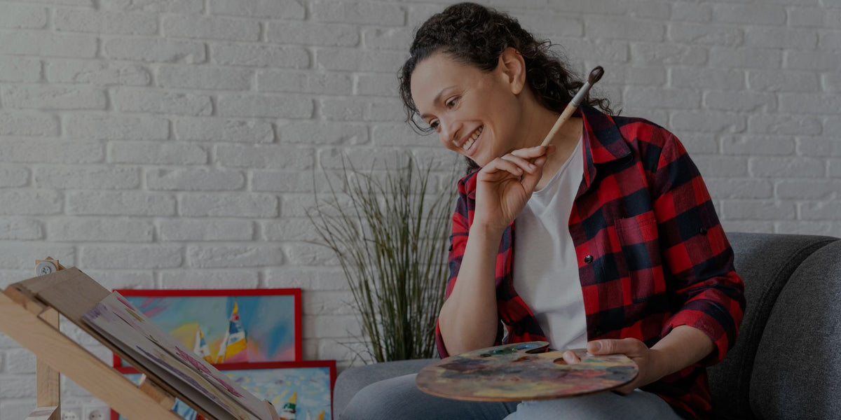 Smiling woman holding paint palette and brush near canvas easel